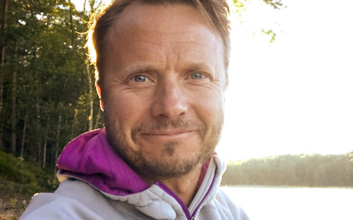 Anders står i skogen vid en sjö och ler vänligt iförd en vit fleece-jacka.