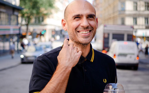 Dimitris har ett brett leende och tittar på något utanför bild. Han har mörk kortärmad tröja och håller i en takeaway-kaffe. I bakgrunden syns en trafikerad gata.