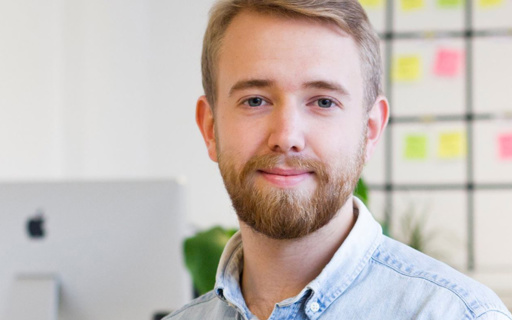 Joel har jeansskjorta och ett välansat kort skägg. I bakgrunden syns en whiteboard med postit-lappar och en datorskärm.