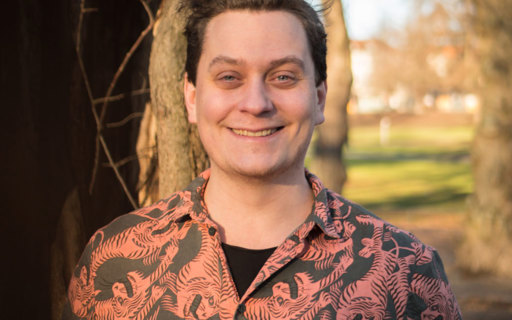 Jonas ler stort bredvid några träd. Han bär en svart skjorta med rött intensivt mönster.