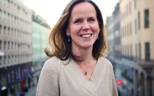 Karin ler ståendes uppe på en bro. Hon har axellångt mörkt hår och bär en beige stickad tröja. I bakgrunden syns en trafikerad gata.