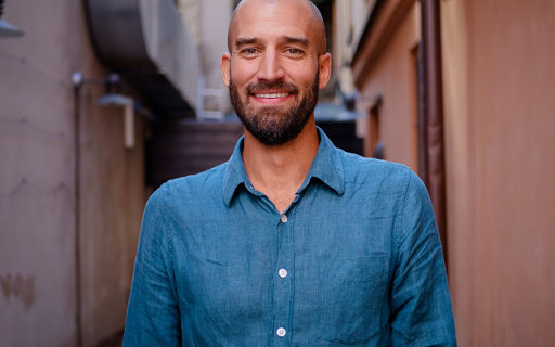 Kristoffer står i en gränd och ler. Han har rakat huvud, mörkt skägg och en blå skjorta.