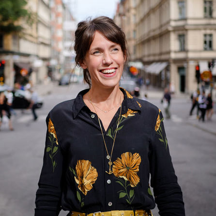 Märta är utomhus på en livfull gata. Hon ler och har en svart blus med stora gula blommor.
