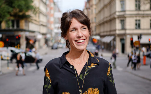 Märta är utomhus på en livfull gata. Hon ler och har en svart blus med stora gula blommor.