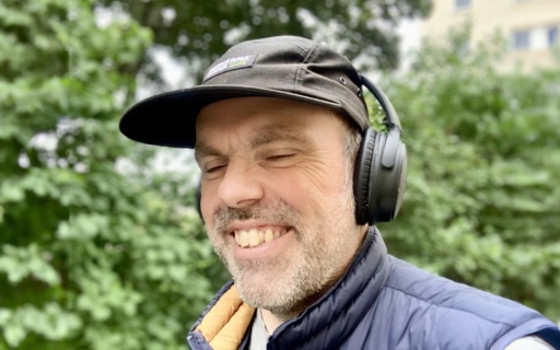 Martin har svart keps, blå väst och hörlurar. Han ler åt något som sägs i hörlurarna. I bakgrunden syns buskar och träd.