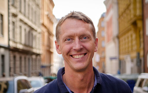 Per ler glatt och bär en mörkblå tröja. Han står mitt i en stadsgata.