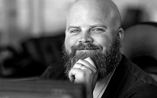 Putte sitter vid sitt skrivbord. Han ler självsäkert och håller upp en hand mot hakan. Hans skägg är stort och lockigt. Svart-vitt foto.